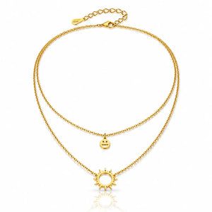 A double-layered gold necklace featuring a delicate chain with a small smiley face charm and a larger sun pendant, displayed on a clean white background with a subtle reflection.