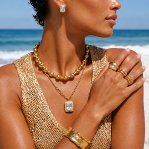 A close-up lifestyle shot of a model on a beach wearing a gold beaded necklace, a gold pendant necklace with a clear rectangular gemstone, a gold ring set with a clear gemstone, a gold cuff bracelet, and a matching gold earring.