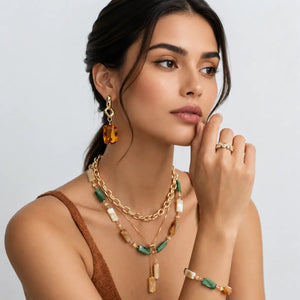 A woman models a coordinated jewelry set featuring a gold-tone layered necklace with green and cream beads, matching drop earrings, and rings, set against a clean light gray background.