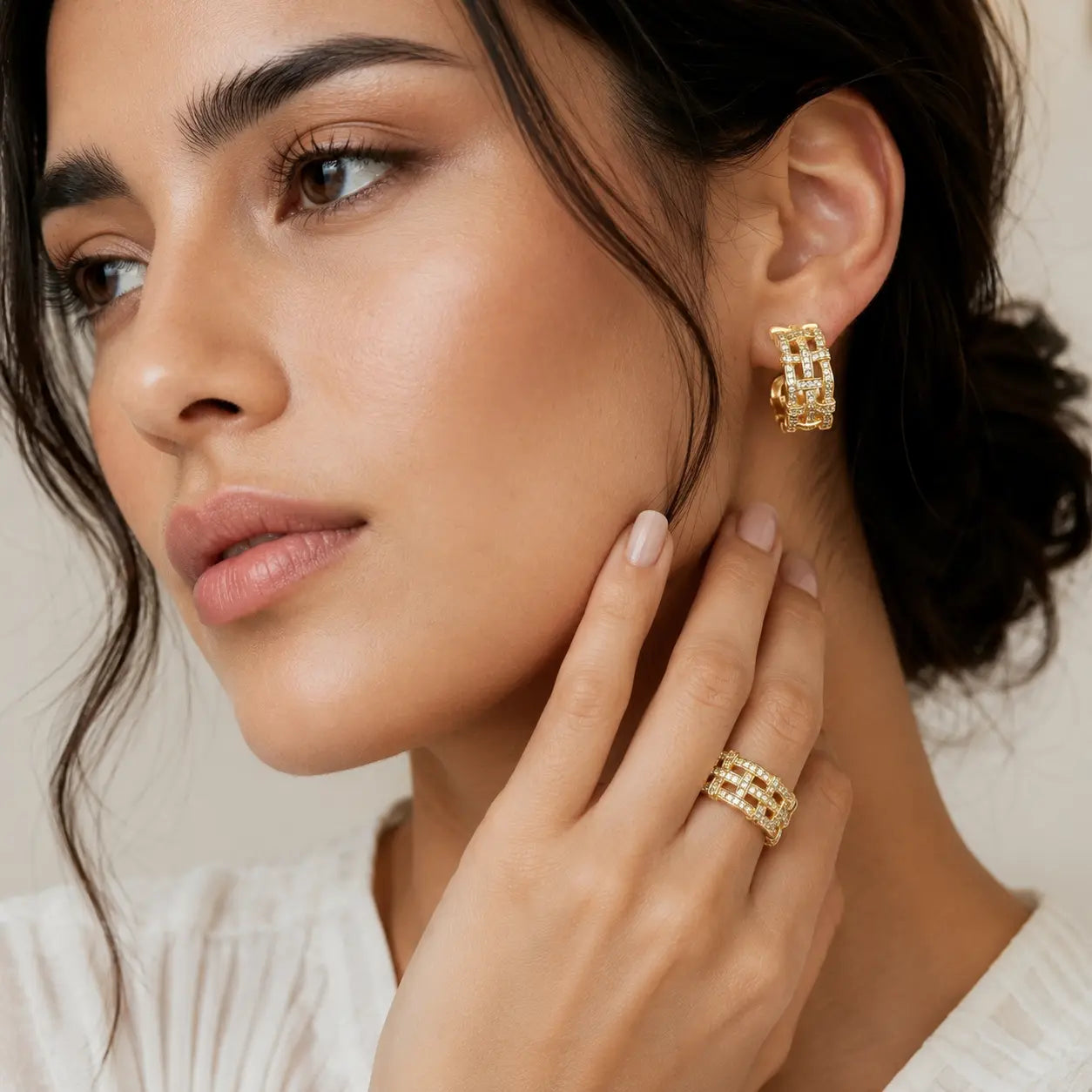 A close-up lifestyle shot of a woman wearing a gold hoop earring and a matching gold ring with small clear stones, set against a soft beige background.