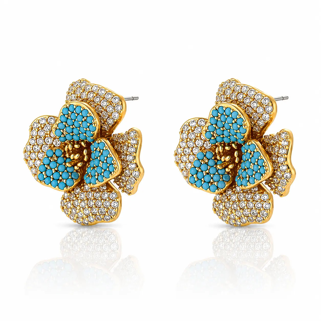 A pair of gold flower-shaped stud earrings embellished with turquoise blue stones and clear pavé crystals, shown on a white background with a soft reflection below.