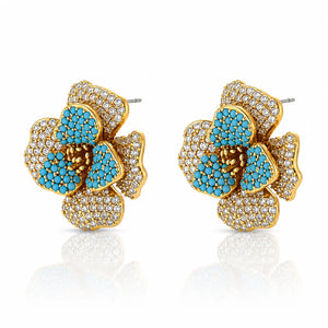 A pair of gold flower-shaped stud earrings embellished with turquoise blue stones and clear pavé crystals, shown on a white background with a soft reflection below.
