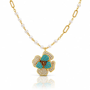 A gold chain necklace featuring white pearls and a gold pendant shaped like a flower, embellished with turquoise blue stones and small clear pavé stones, shown on a white background with a soft reflection.