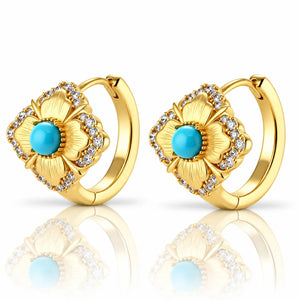 A pair of gold huggie hoop earrings featuring a flower design with a central turquoise blue stone and clear rhinestone accents, shown on a reflective white surface.