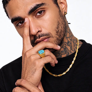 A close-up lifestyle shot of a man wearing a chunky gold chain necklace and a gold signet ring with a turquoise stone, against a clean white background.