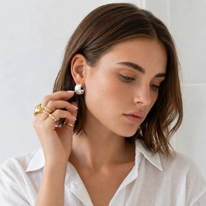 A woman with brown hair wearing a white collared shirt and gold rings, looking down while wearing a small white pearl-like hoop earring.