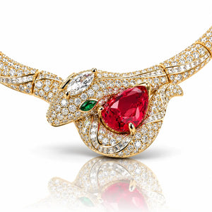 A gold-toned snake head necklace embellished with pavé-set clear crystals, a large teardrop-shaped red gemstone, and smaller green and clear accent stones, shown on a white background with a soft reflection below.