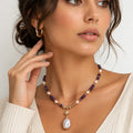 A close-up of a woman wearing a beaded necklace with a baroque pearl pendant and a gold-toned starburst charm, paired with a matching gold hoop earring.