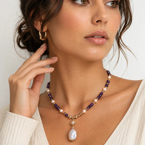 A close-up of a woman wearing a beaded necklace with a baroque pearl pendant and a gold-toned starburst charm, paired with a matching gold hoop earring.