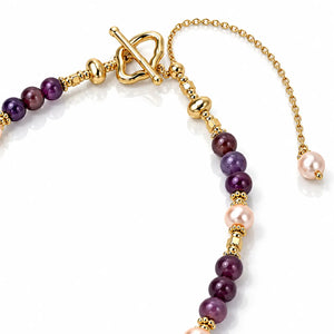 A close-up of a beaded strand jewelry piece featuring dark purple marbled beads, light pink freshwater pearls, and polished gold-tone spacer beads and hardware on a white background.