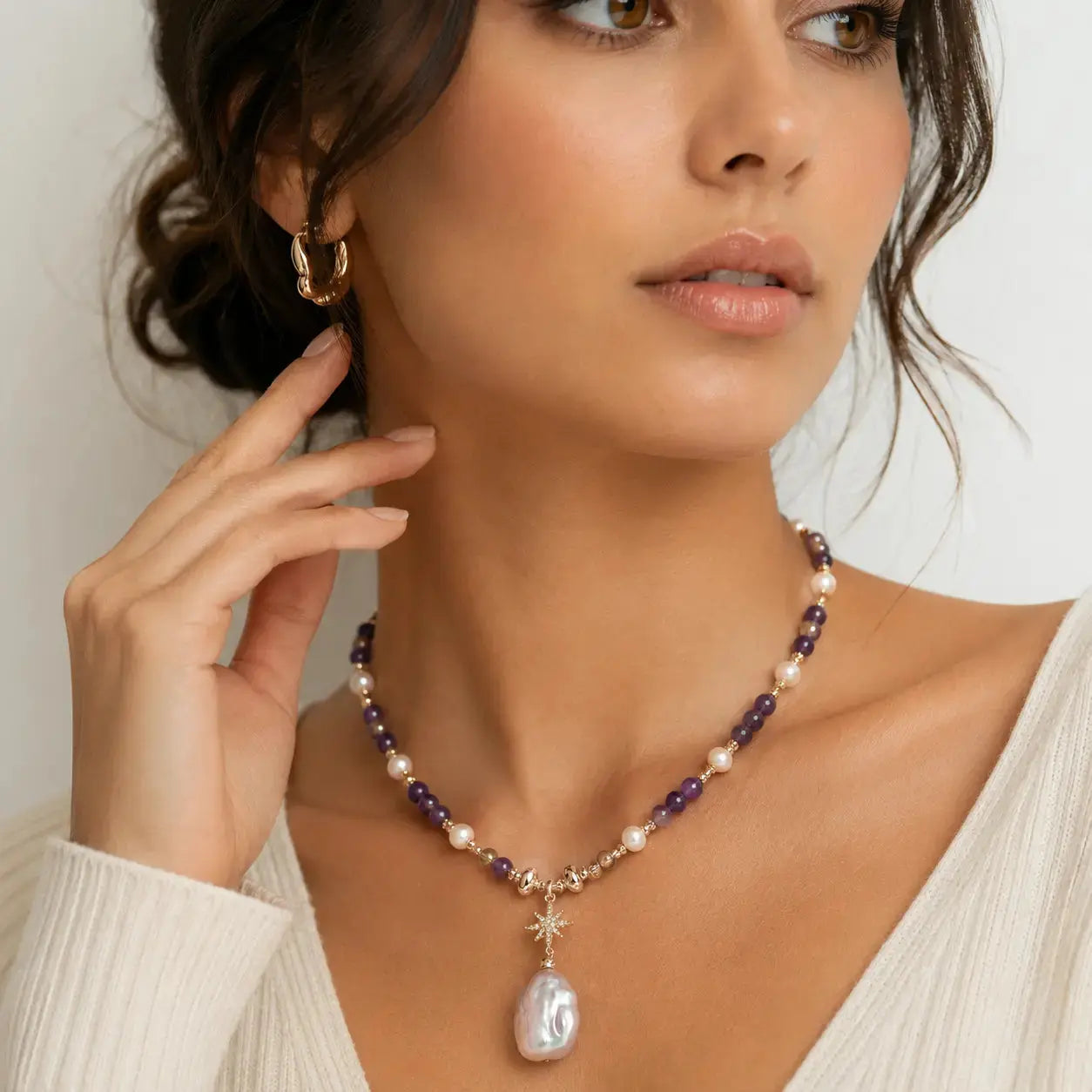 A close-up of a woman wearing a beaded necklace with a large teardrop pearl pendant, purple beads, white pearls, and a star charm, paired with a gold hoop earring.