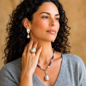 A woman with curly dark hair models a gold and pearl jewelry set, including a drop earring, a ring, and a beaded necklace with a large teardrop pearl pendant, against a warm beige background.