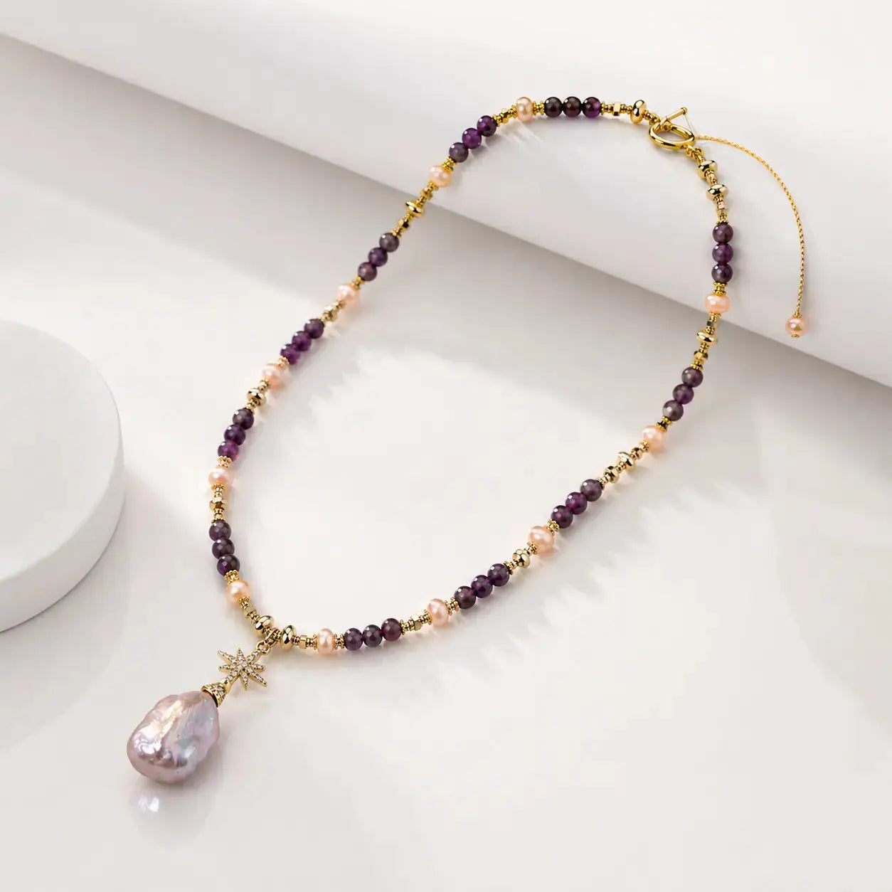 A beaded necklace with purple and peach pearls, gold spacer beads, and a large teardrop-shaped iridescent pearl pendant, displayed on a clean white surface.
