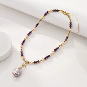 A beaded necklace with purple and peach pearls, gold spacer beads, and a large teardrop-shaped iridescent pearl pendant, displayed on a clean white surface.