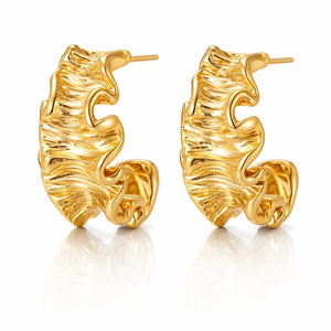 A pair of gold huggie hoop earrings with a textured, organic wavy surface, displayed on a clean white background with a soft reflection below.