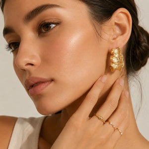 A close-up lifestyle shot of a model wearing a chunky, organic-shaped textured gold hoop earring and delicate gold rings on her fingers.