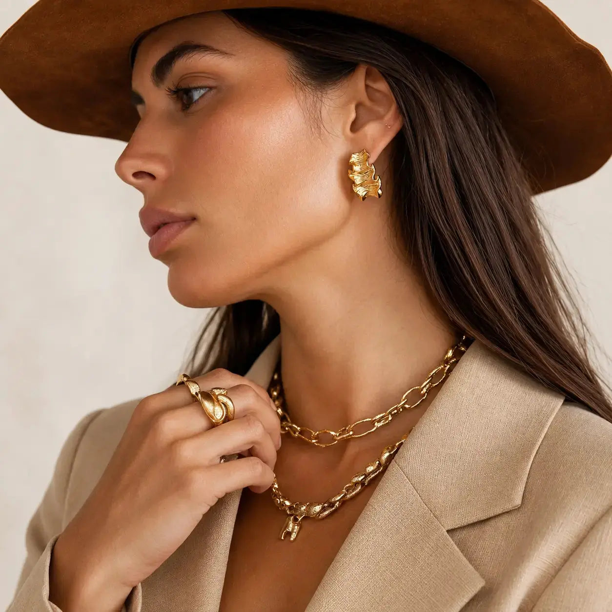 A close-up side profile of a woman wearing a brown wide-brim hat, beige blazer, and layered gold jewelry including a textured gold earring, double layered chain necklaces, and stacked gold rings.