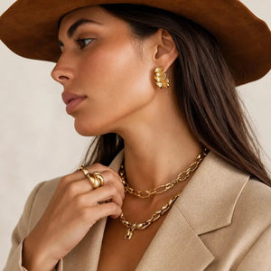 A close-up side profile of a woman wearing a brown wide-brim hat, beige blazer, and layered gold jewelry including a textured gold earring, double layered chain necklaces, and stacked gold rings.
