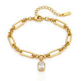 A gold paperclip chain bracelet with a rectangular clear gemstone pendant, displayed on a clean white background with a subtle reflection.