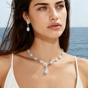 A woman outdoors by the sea wearing a sparkling silver-toned necklace with clear gemstones and matching drop earrings.
